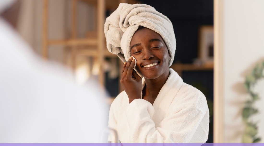 Smiling woman in a white bathrobe and towel applying skincare to her face in a warm, cozy bathroom setting.